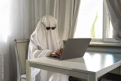 White bed sheet ghost costume typing on a laptop in a grey monochrome spooky home office