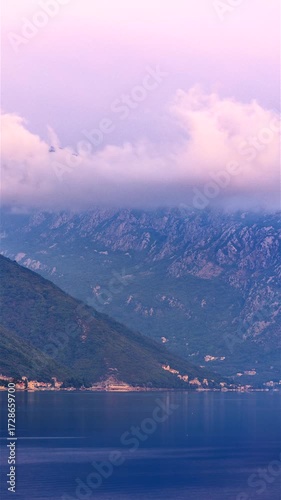 Pre-dawn view of Kotor Bay, Montenegro, with pastel pink and blue sky, low clouds over mountains, and calm reflective water before sunrise. Time lapse