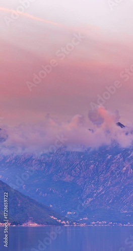 Pre-dawn view of Kotor Bay, Montenegro, with pastel pink and blue sky, low clouds over mountains, and calm reflective water before sunrise. Time lapse