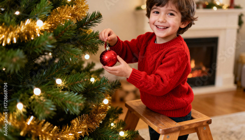 Cute smiling child hanging red ball ornament on Christmas tree. Happy boy in red sweater enjoying festive holiday season. Winter celebration and family tradition at home concept.