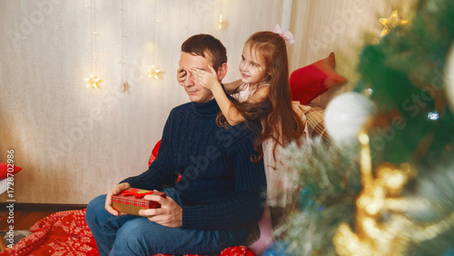 A little girl closes her father's eyes as she surprises him with a gift box on the Christmas. Near the Christmas tree at home