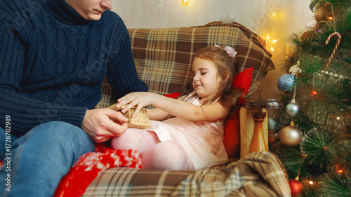 Dad is giving a gift box to his beautiful little daughter. Near the Christmas tree at home