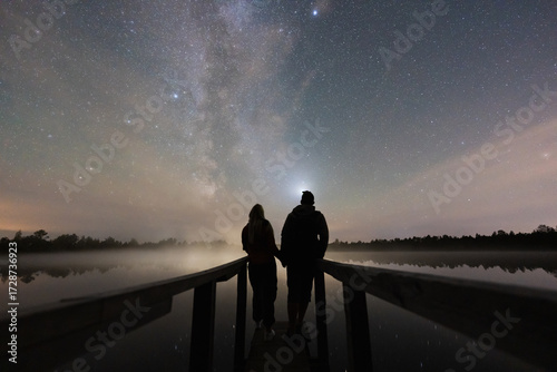 A romantic couple on a wooden pier enjoys the magical starry night sky with the Milky Way in a tranquil Estonian bog.
