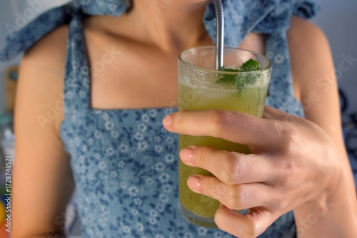 Woman drinking from metal straw green kiwi lemonade in glass with mint leaves on kitchen at home. Recipe cuisine professional culinary. Prepare delicious fruit dessert homemade beverage drink.