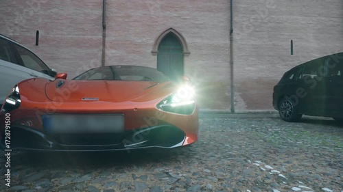 Sleek orange sports car parked on the street with switched on headlights against background of red brick building, exclusive collection of expensive automobiles with shiny design, sports cars racing