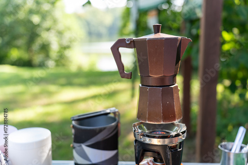 Obraz na płótnie Brown moka pot coffee maker brewing on portable gas burner with white cup and camping equipment on outdoor table