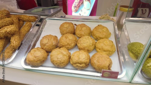 Assorted fried pastries and cream puffs displayed on metal trays in a cafe.