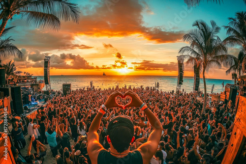 A large crowd gathers on the beach to watch a stunning sunset while enjoying music and celebrating together during a festival
