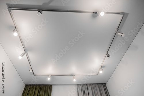 A fragment of a modern interior, shot from below, shows a rectangular track lighting system with nine white spotlights installed on the white ceiling.

