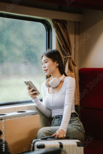 Smiling young woman traveler with suitcase and headphones around her neck holding a smartphone at the train station. Free Download Smiling young woman traveler with suitcase and headphones around her neck holding a smartphone at the train station. - GFXLoad