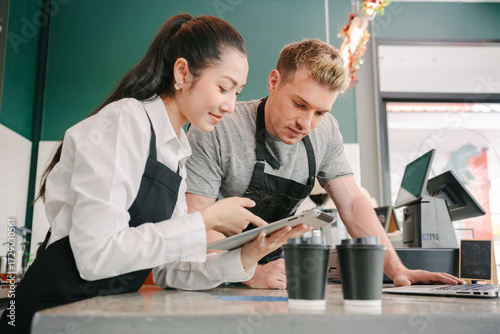 Two baristas, a man and a woman in black aprons, work together behind counter. The man is looking at a laptop, and the woman is checking a tablet while standing next to a couple of to-go coffee cups.