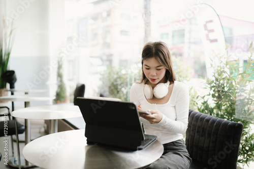 Young Asian woman wearing headphones around her neck, sitting at a coffee shop with laptop and smartphone, enjoying modern digital lifestyle. Free Download Young Asian woman wearing headphones around her neck, sitting at a coffee shop with laptop and smartphone, enjoying modern digital lifestyle. - GFXLoad