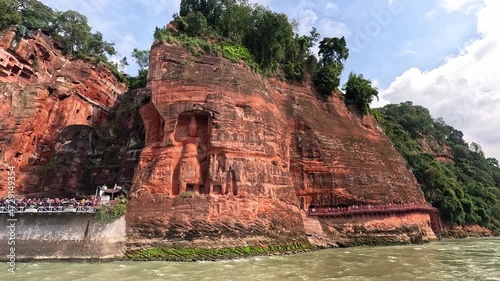 Slow approach from river of Leshan Giant Buddha framed by red cliffs and green trees, showing monumental scale of mountain carving
