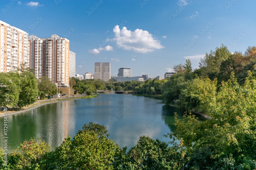 Fototapeta premium This serene landscape features a tranquil lake reflecting the blue sky, with lush trees along the banks and contemporary buildings in the background, showcasing urban nature harmony
