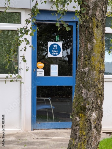 Blue office exterior door marked with a fire exit sign that has a chair on the inside blocking the safe exit route in the case of a fire breaching all health and safety laws and fire regulations.