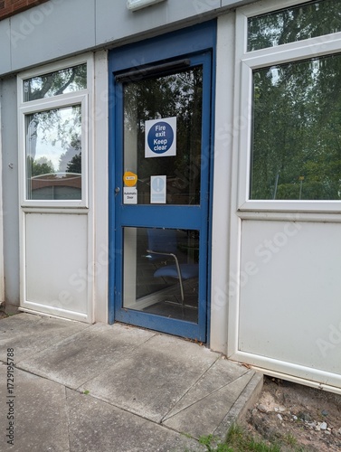 Blue office exterior door marked with a fire exit sign that has a chair on the inside blocking the safe exit route in the case of a fire breaching all health and safety laws and fire regulations.