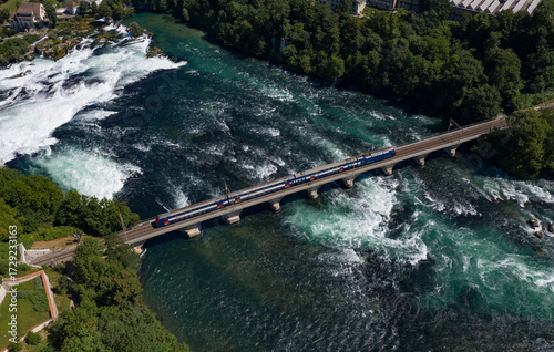 Aerial view of a train crossing a bridge over the churning, turquoise Rhine River, with the powerful Rhine Falls cascading nearby, Filisur, Switzerland.