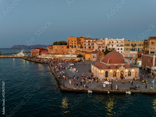 Aerial view of the Kioutsouk Hassan Mosque with its prominent dome glows warmly amidst the bustling harbor, a vibrant contrast to the deep blue sea, Chania, Chania, Greece.