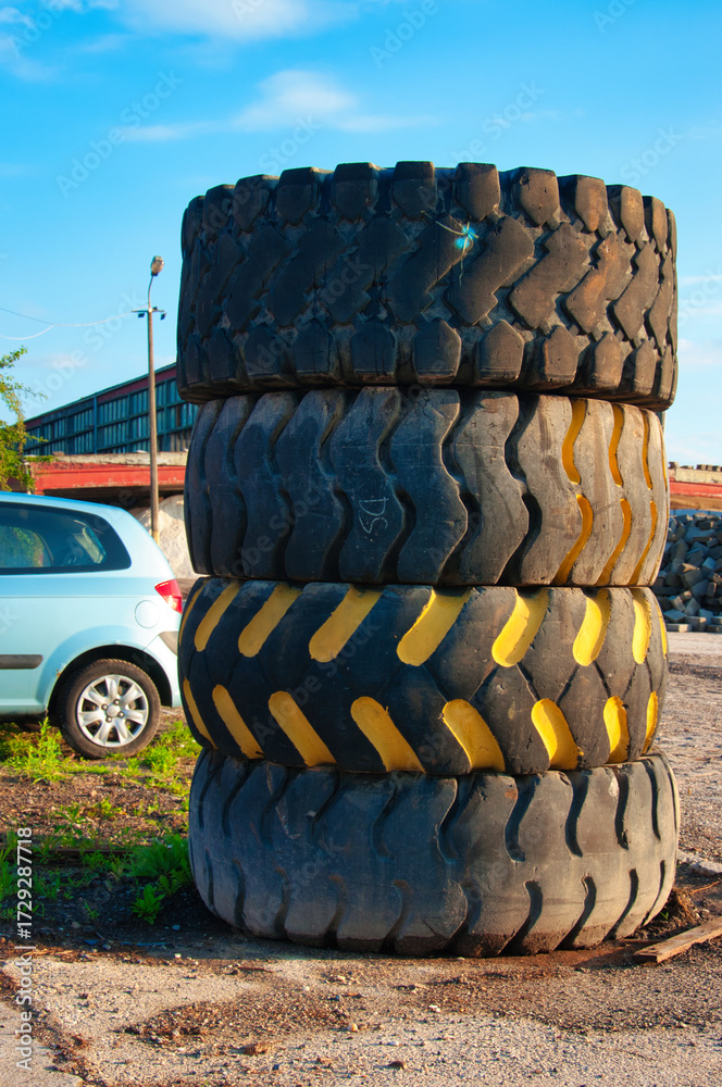 Fototapeta premium Wysoki stos zużytych potężnych opon leży na zakurzonym podwórku przemysłowym. W pobliżu zaparkowany jest jasnoniebieski samochódo z małą oponą oświetlony promieniami słońca.