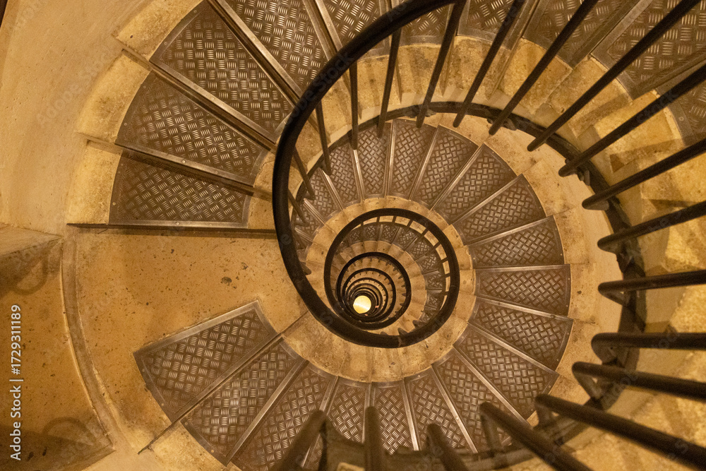 Fototapeta premium Spiral staircase viewed from above, showcasing intricate metal railings and textured steps, creating a captivating architectural design with depth and perspective