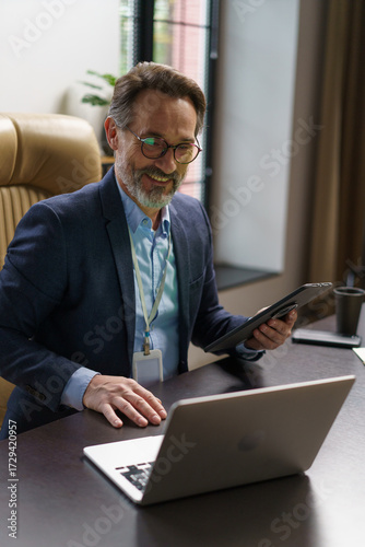 A confident man in business attire smiles as he participates in a virtual meeting, holding a tablet in one hand while working on a laptop at his sleek office desk