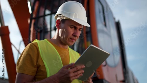 Inspecting crane and machinery with tablet construction worker wearing helmet and vest checks tablet on site near orange crane while operator inspects for safety and maintenance planning process