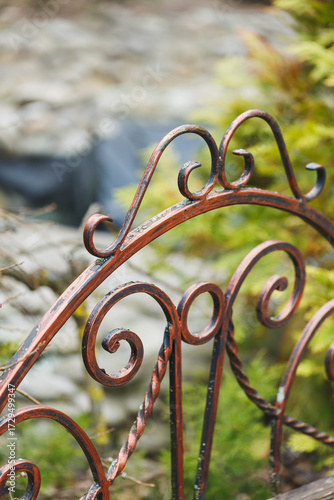 Intricate red metal gate adorned with swirling designs opens to a lush garden surrounded by natural beauty