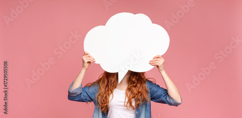 Unrecognizable redhead female holding an empty speech bubble in front of her head, pink studio background, panorama with empty space