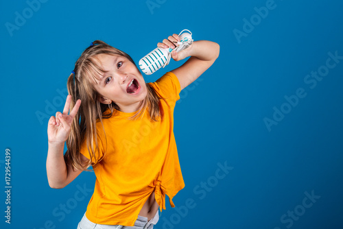 Cute european blonde girl with toy microphone singing against blue background. Rock star concept.