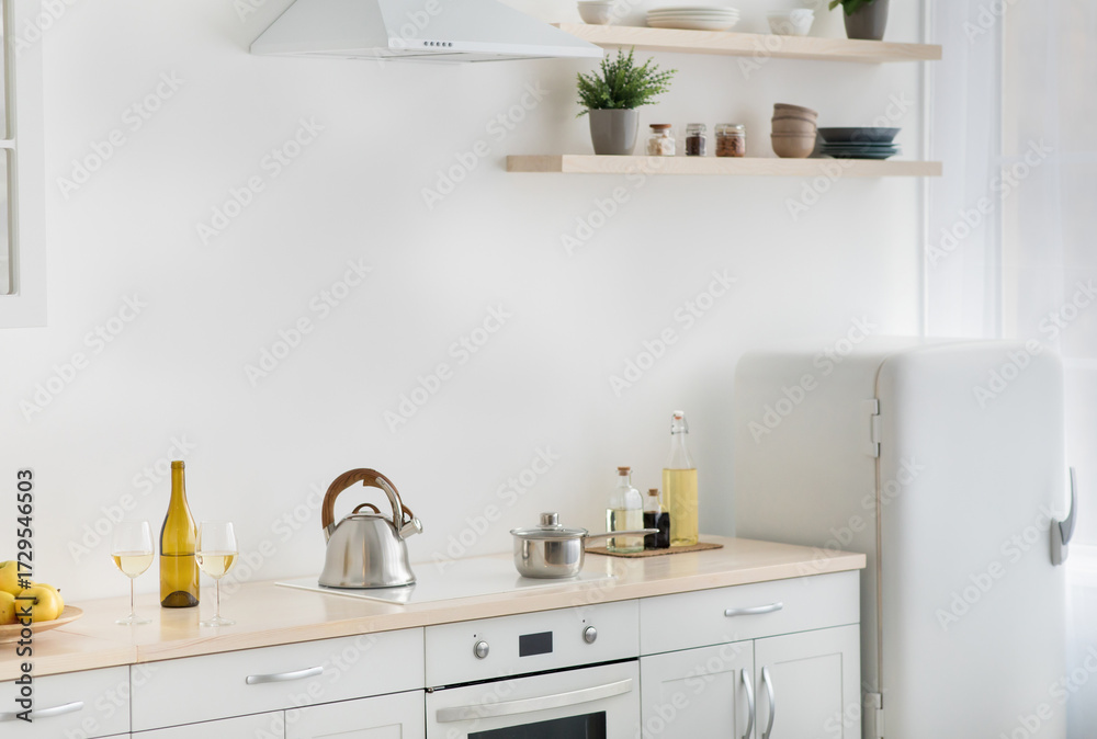 Fototapeta premium Rent flat, real estate, ready for cooking, domestic culinary, home healthy eat. White kitchen, refrigerator, stove with kettle, plate with apples, shelves with potted plants and utensils, light wall
