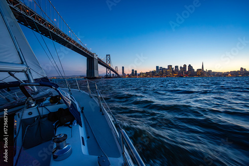 Photography A sailboat cruises on the water at dusk, passing under the Bay Bridge with the San Francisco skyline in the background