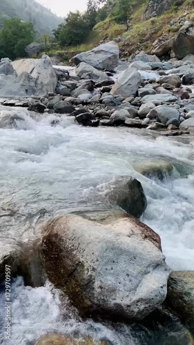 A powerful mountain stream flowing through large rocks and boulders, creating white foamy rapids. The crystal-clear water rushes with energy, showcasing the beauty of untouched nature in a rocky river