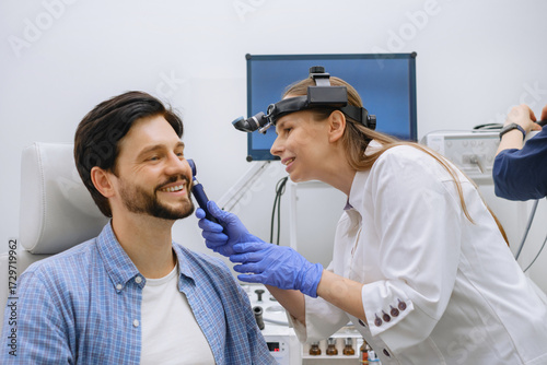 ENT doctor examining male patient with otoscope in modern clinic. Otolaryngology specialist in gloves and headlamp during ear examination.