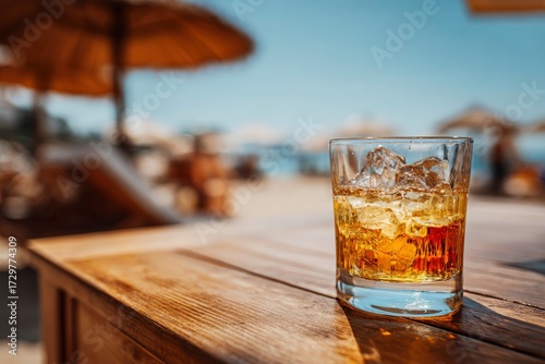 Glass of amber liquid with ice sits on a wooden surface a beach setting blurs in the background