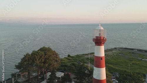 Aerial Red white lighthouse on Black Sea coast at sunset, Poti, Georgia. Calm sea, swimmers, cars, coastal park, navigation landmark, maritime safety, travel icon, summer evening light.