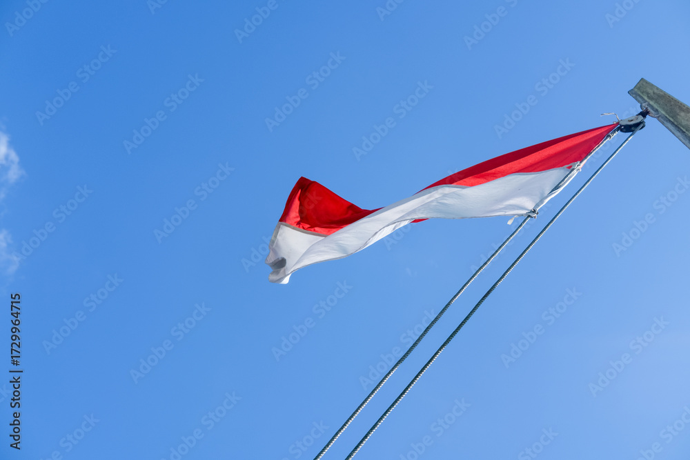 Fototapeta premium Indonesian flag waving proudly on a tall wooden pole against a clear blue sky, symbolizing independence, unity, and national pride with its red and white colors standing out brightly.