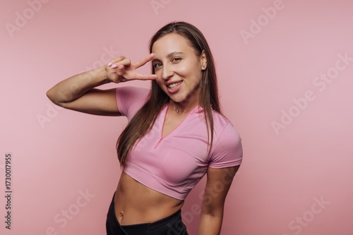 Smiling woman in pink top making peace sign on pink background.