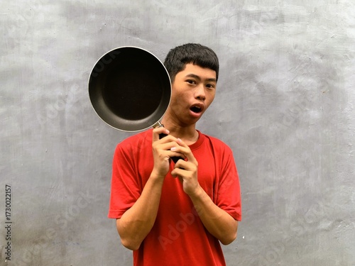 A southeast asian teenager with shocked and startled expression in a red t-shirt carries a frying pan