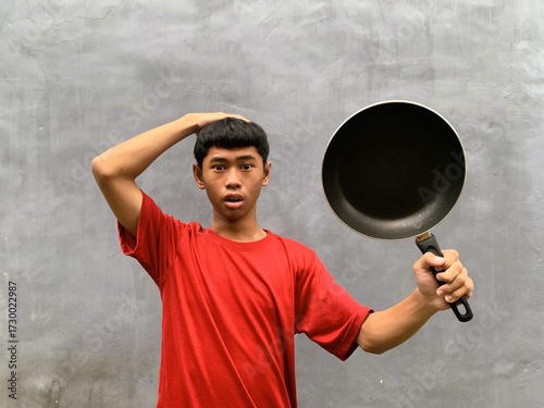 A southeast asian teenager with shocked and startled expression in a red t-shirt carries a frying pan