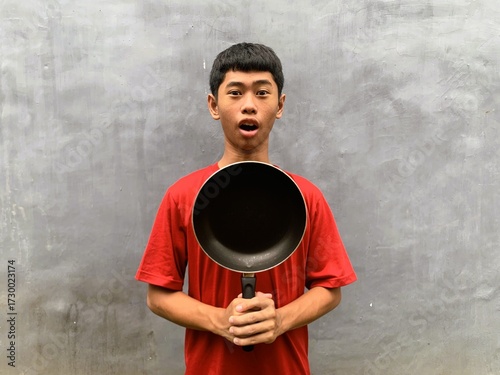 A southeast asian teenager with shocked and startled expression in a red t-shirt carries a frying pan