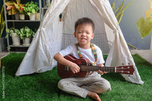 Cheerful young Asian boy sitting on artificial grass in backyard playing ukulele with colorful strap smiling happily in front of white tent surrounded by green plants and natural light