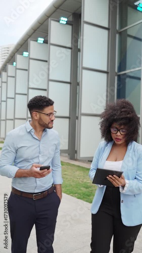 Vertical video of two diverse Business colleagues walking and discussing work using tablet and phone outside office