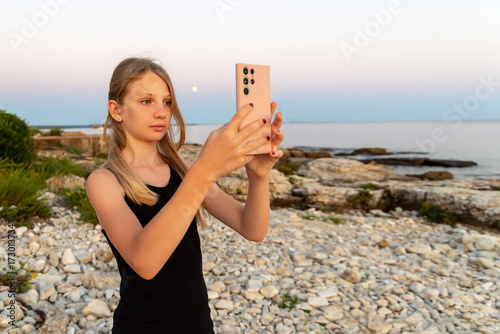 Teen girl taking photo with phone at rocky beach. A young teenage girl uses a smartphone to capture a photo at a rocky coastal area during sunset or moonrise with calm sea background.