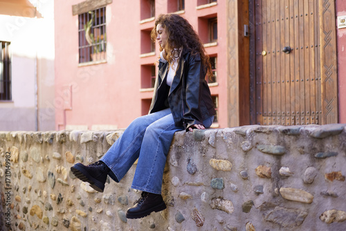 A young woman sits on a stone wall in a trendy outfit, showcasing her style and confidence