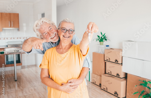 Cheerful senior couple hug moving into a new home, surrounded by moving boxes in an empty room. The woman holding house keys, excited for their new beginning, real estate, property concept
