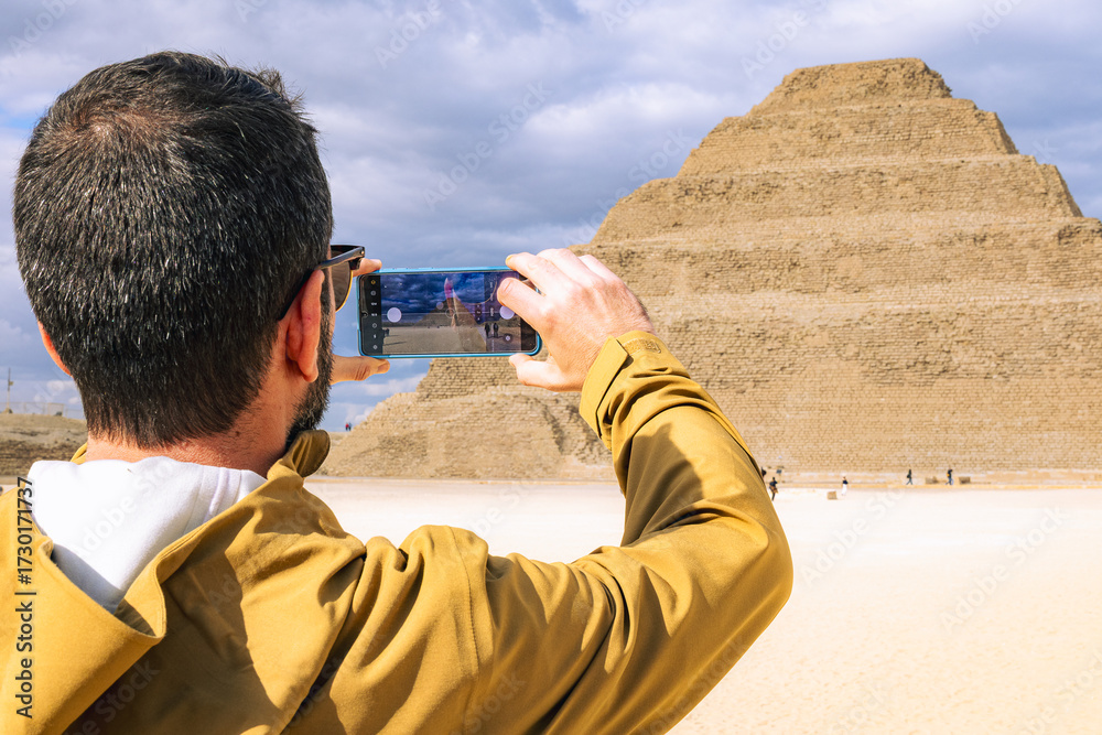 Φωτογραφία A tourist uses a smartphone to photograph the ancient Step Pyramid of Djoser in Saqqara, Egypt, capturing a moment of modern travel and ancient history