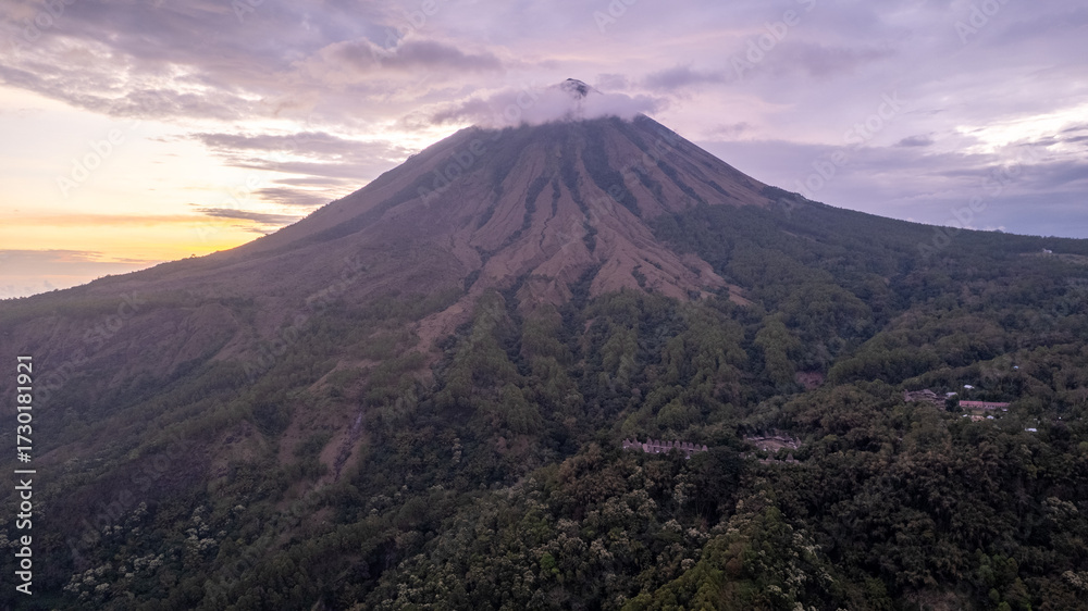 Fototapeta premium A breathtaking view of Mount Inerie in Flores, Indonesia, during sunset. The volcanic peak rises steeply with clouds at the summit, surrounded by lush green forest and dramatic sky. 