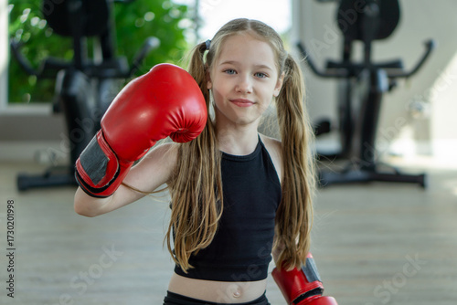 A young girl is posing with oversized red boxing gloves in a well-lit gym. She exudes confidence and joy while preparing for her boxing practice.