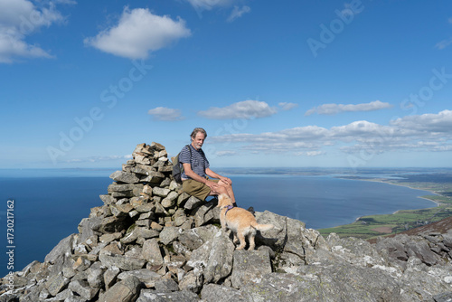 Active senior man sitting on a mountain summit cairn with a smile while stroking a golden dog under a blue sky with cloud and views over the ocean.