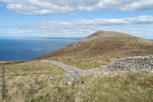 Stone wall leads to the summit peak of Gyrn Goch mountain, Gwynedd, Wales, UK, with views over the sea under a blue sky with cloud.
